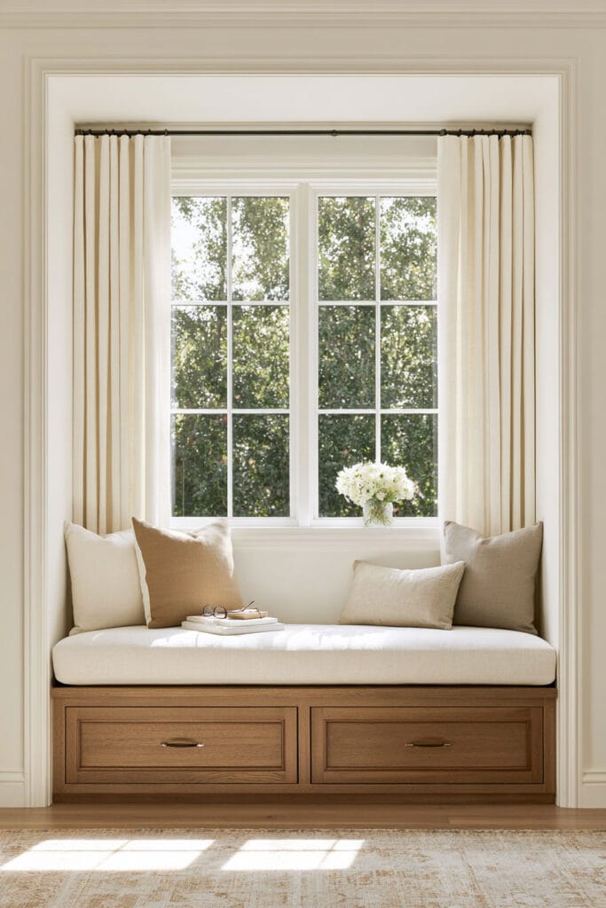 Light and airy window reading nook with a built-in wooden bench, cream cushions, beige pillows, floor-to-ceiling ivory curtains, storage drawers, and natural sunlight.